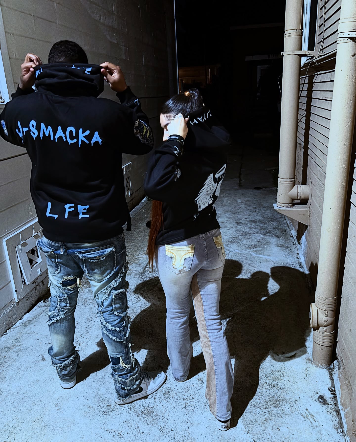 Two people from behind in an alleyway, one wearing a J-SMACKA LIFE hoodie and one wearing a ZARAC LANE hoodie